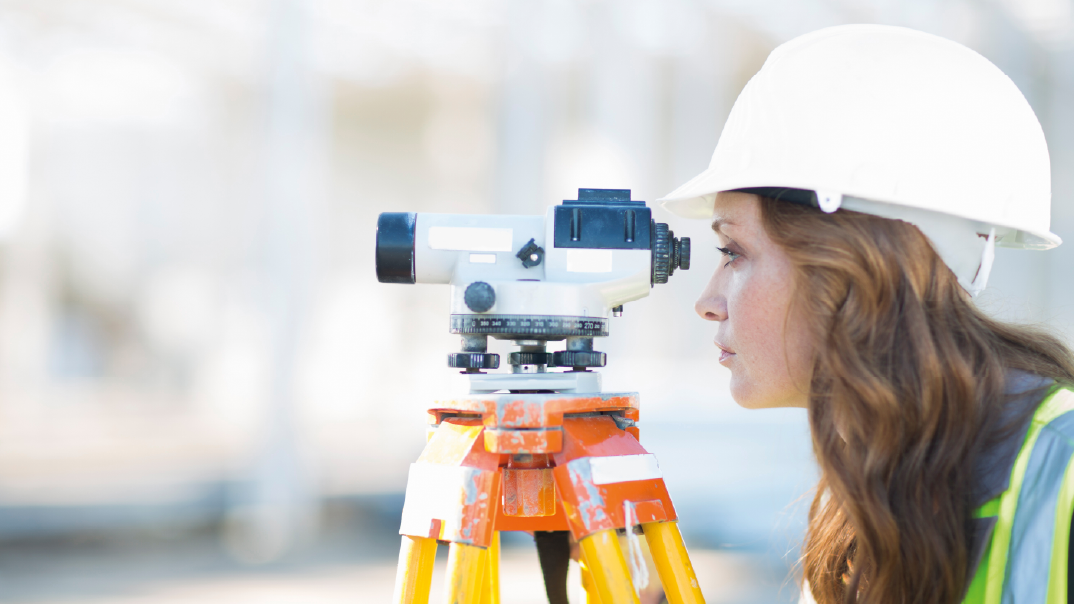 A woman in a white hardhat and a safety vest looks into a laser level measuring tool on a tripod.