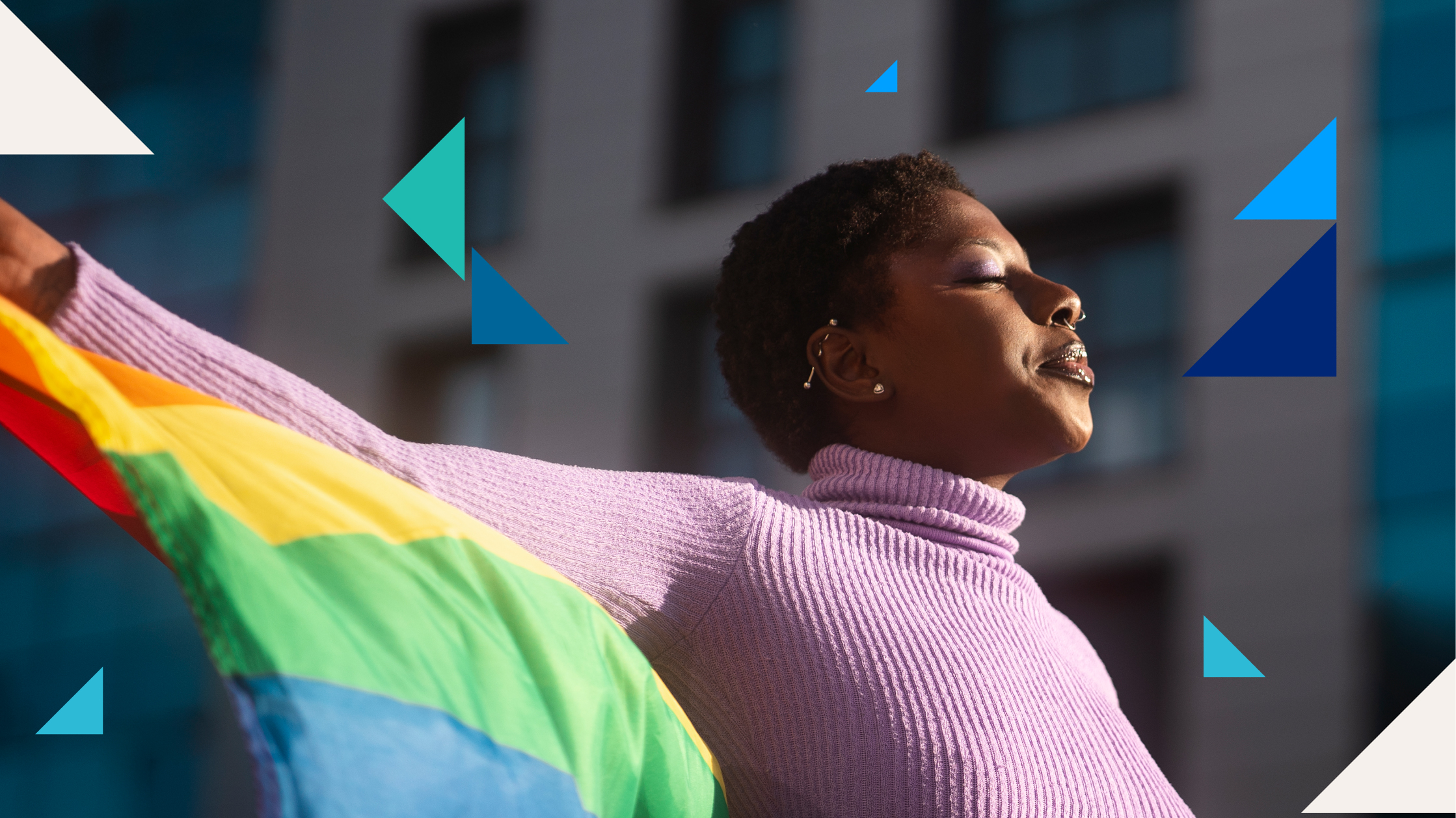 A woman in a purple sweater and closed eyes holds a rainbow Pride flag behind her as the sun shines on her.