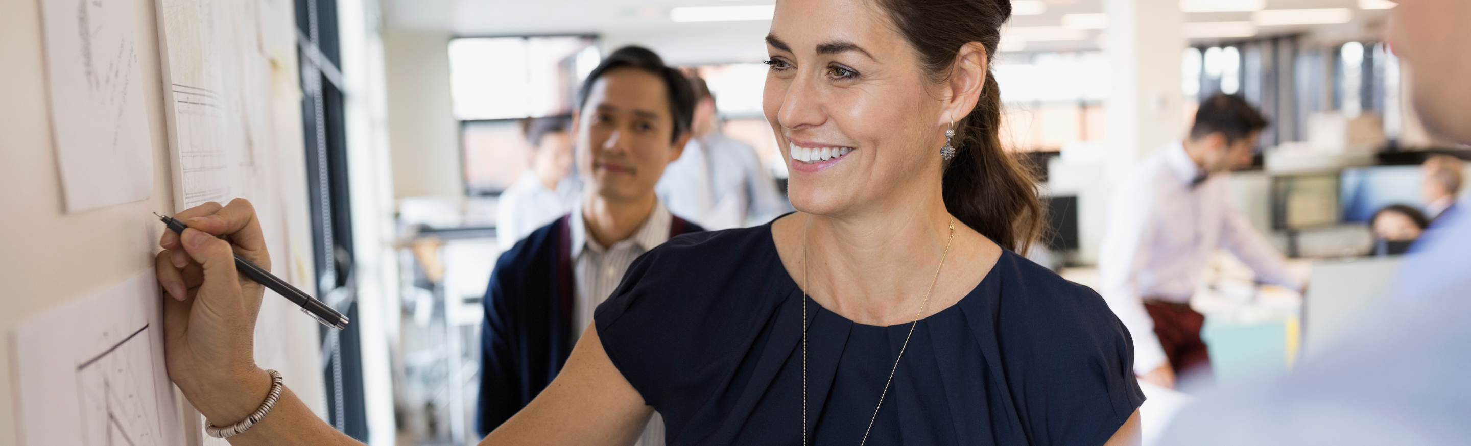 Employee engagement strategies guide cover: A woman in a dark blue blouse smiles brightly as she writes on a whiteboard while a man looks on behind her in an office setting.