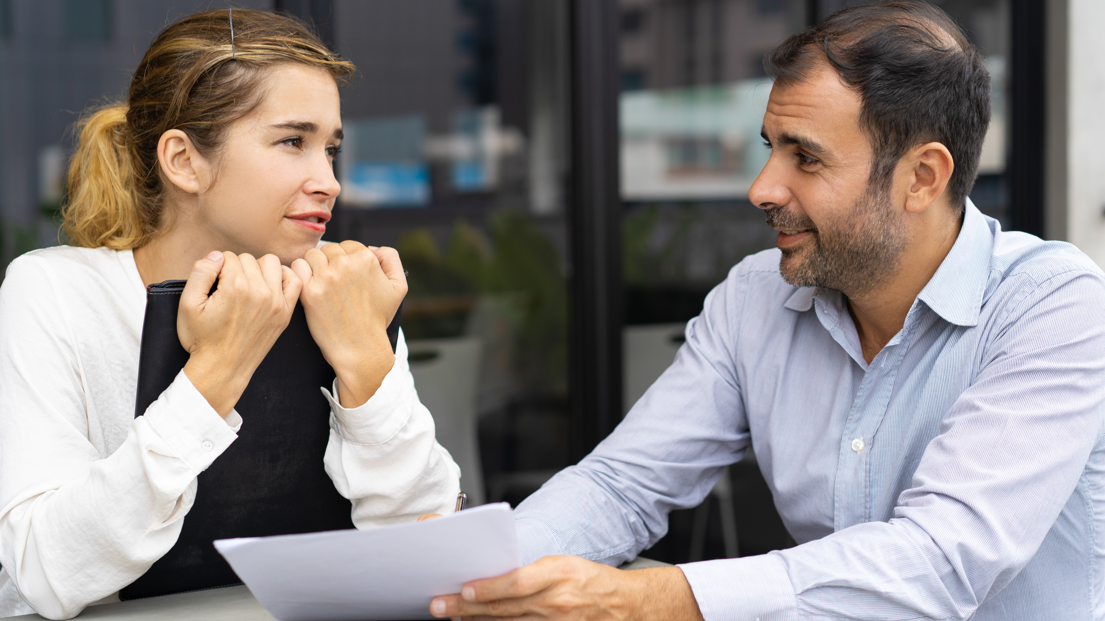 A woman looks at a man. She holds a binder in her arms, the man is holding a piece of paper at her.