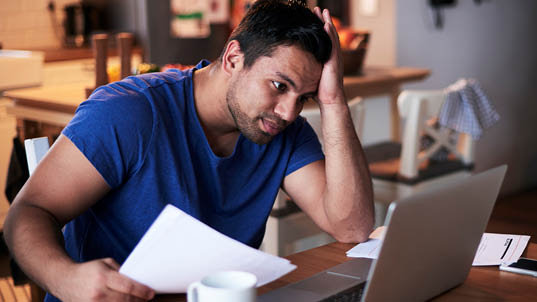 A man in a blue shirt holds his head while he looks at a laptop. He looks troubled.