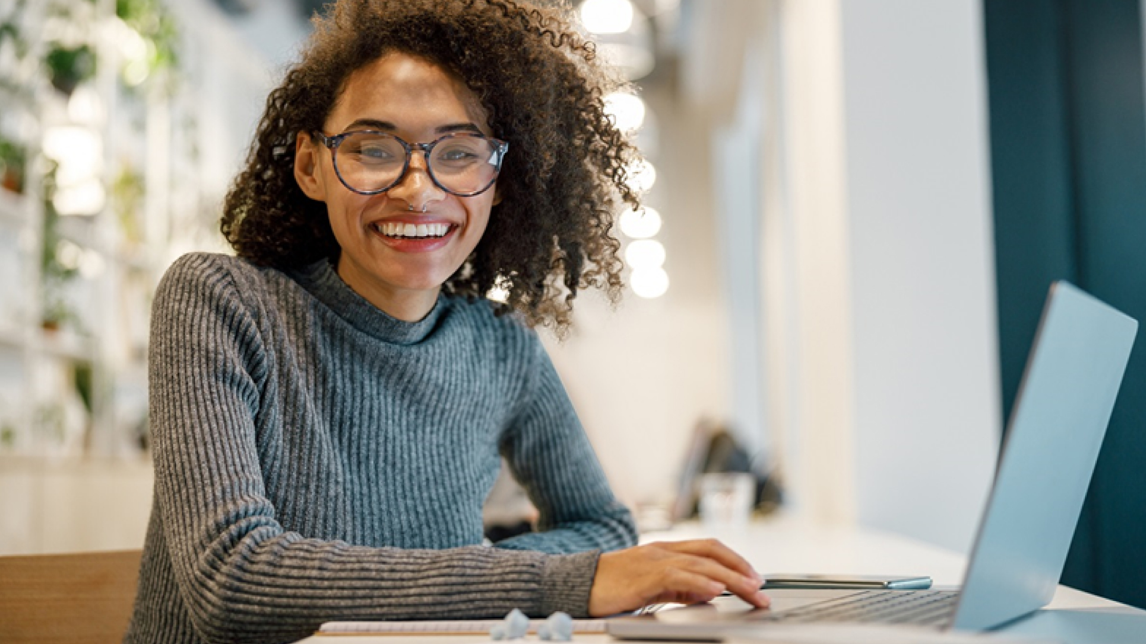 A woman in a gary sweater and big, brown glasses smiles at the camera. In front of her is a grey laptop.