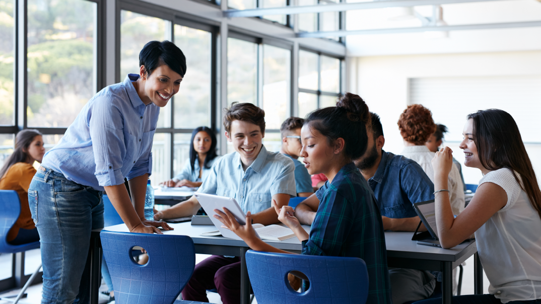 A group of students smile around a table while showing their teacher something in their tablet.