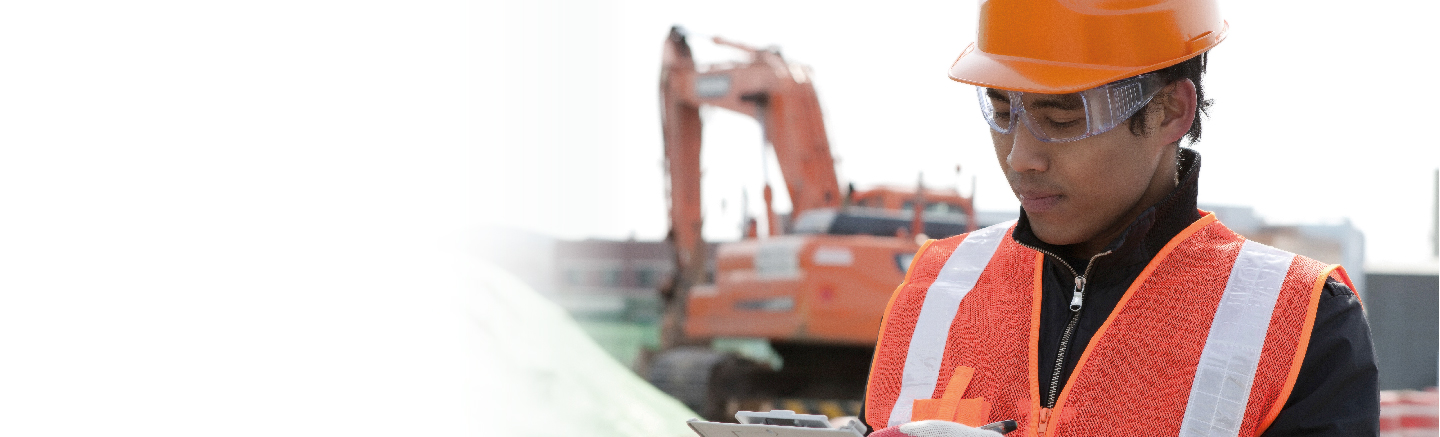 A man wearing an orange hard hat, security vest and goggles. Behind him is an orange excavator in the distance.