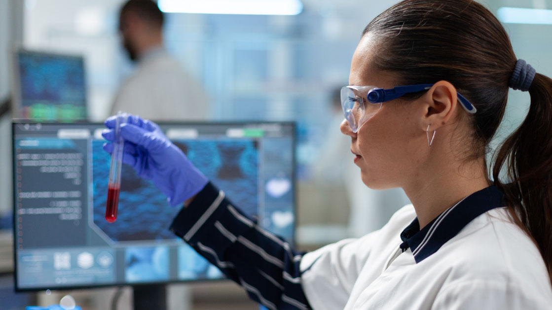 A woman in a laboratory stares at a blood vial. She is wearing protective goggles and a striped navy blue shirt.